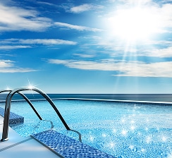 Piscine à débordement luxe vue mer et soleil éclatant Escalier métallique entrant dans une piscine à débordement carrelée en bleu, avec le soleil éclatant et l'océan à l'horizon.