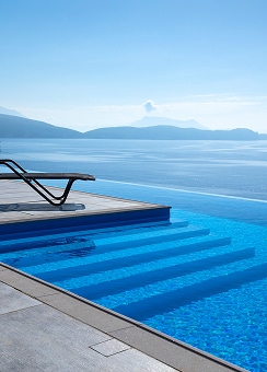Piscine à débordement vue mer : luxe et sérénité Piscine à débordement bleue aux marches immergées, avec une chaise longue noire face à une mer calme et des montagnes lointaines sous un ciel clair.