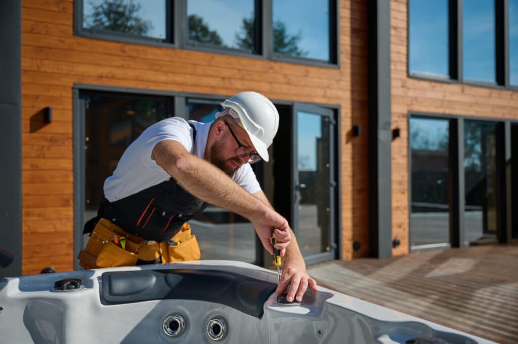 Technicien barbu en casque blanc visse un bouchon sur un jacuzzi extérieur, devant une maison moderne à bardage bois.